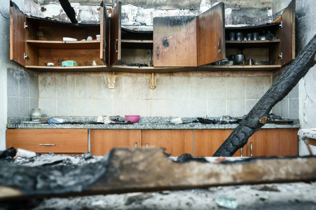 Fire-damaged kitchen with soot-stained cabinets, charred surfaces, and debris across benchtops and flooring