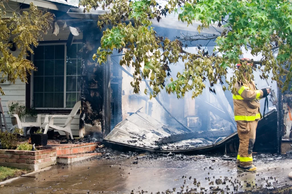 Firefighter inspecting a house after fire damage, with smoke, debris, and a collapsed garage structure visible
